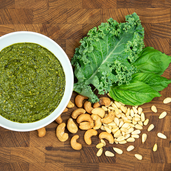 A bowl of green pesto surrounded by fresh basil leaves, kale, cashews, and pine nuts on a dark wood cutting board.